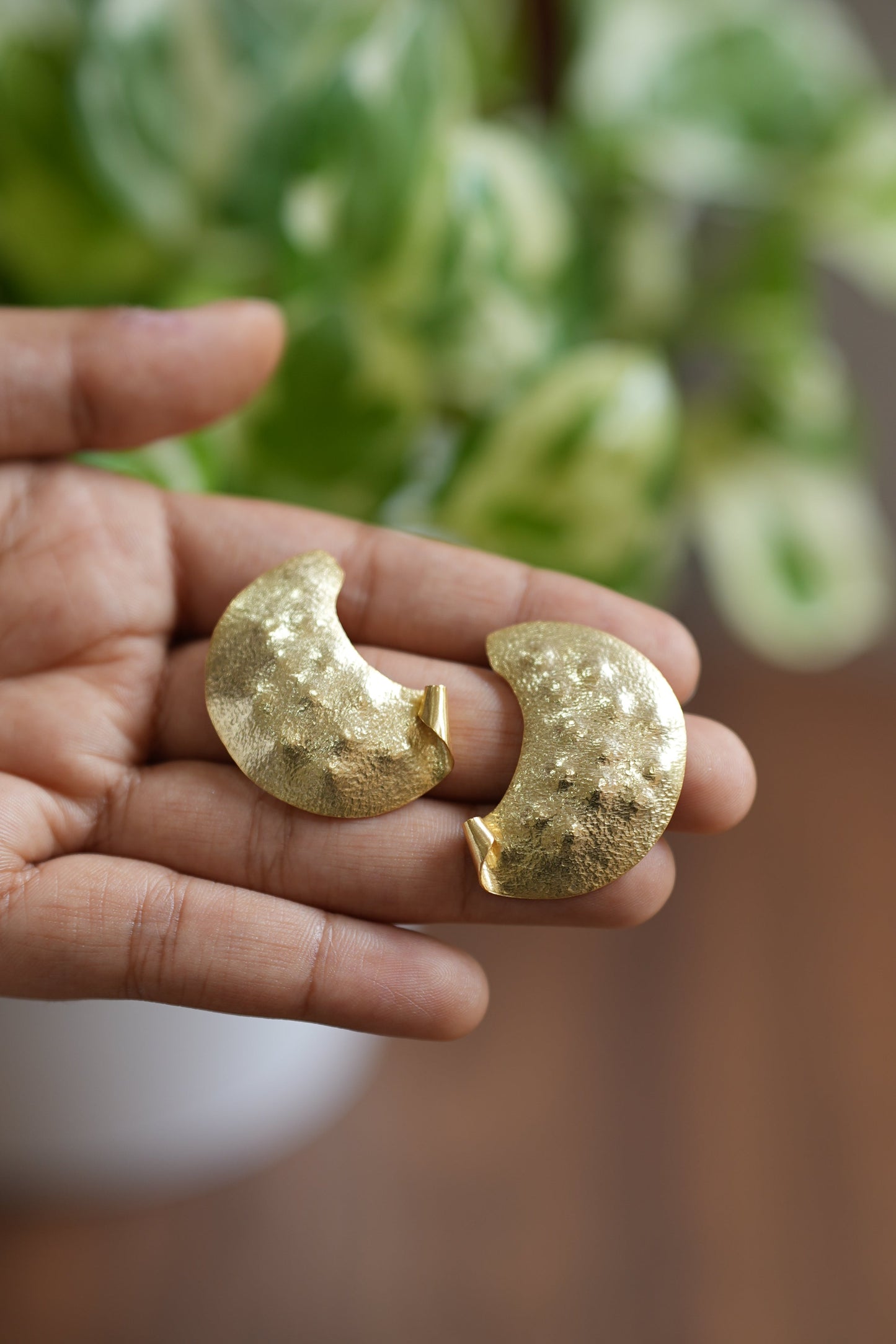 Gold earrings held in a hand with a blurred plant and wooden surface in the background