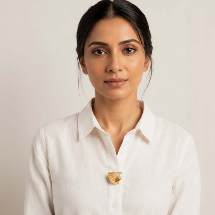A woman with dark hair pulled back wears a white long-sleeve button-up shirt with a small gold geometric pendant at the center of the chest, set against a plain light beige background.