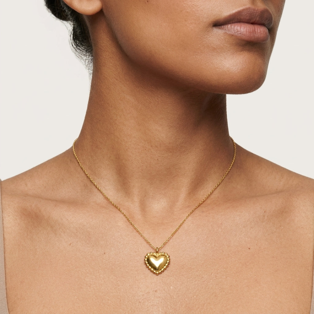 A close-up of a model wearing a delicate gold chain necklace with a small heart-shaped pendant, shown against a neutral light beige background.