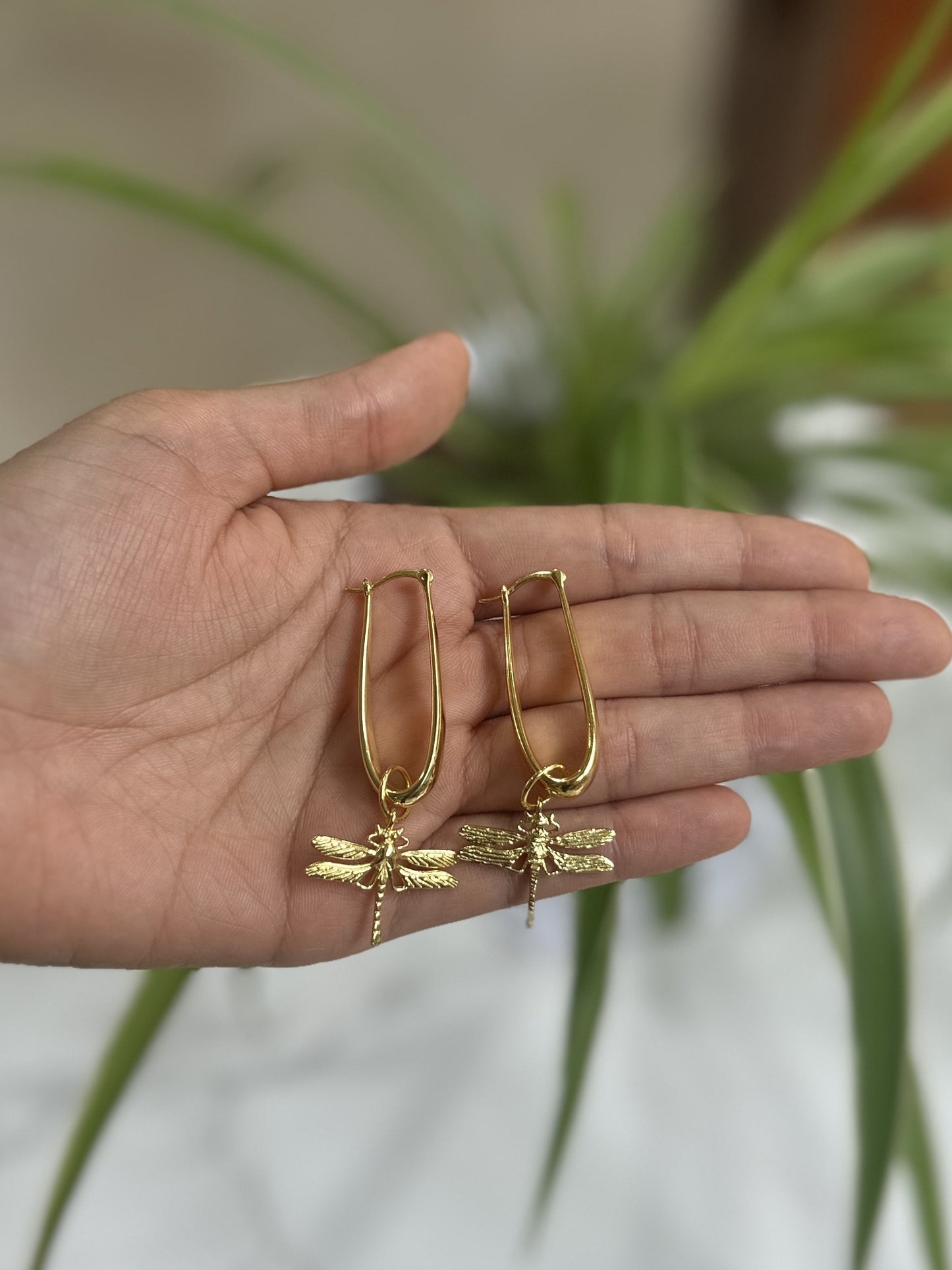 Gold dragonfly earrings held in a hand with a plant in the background