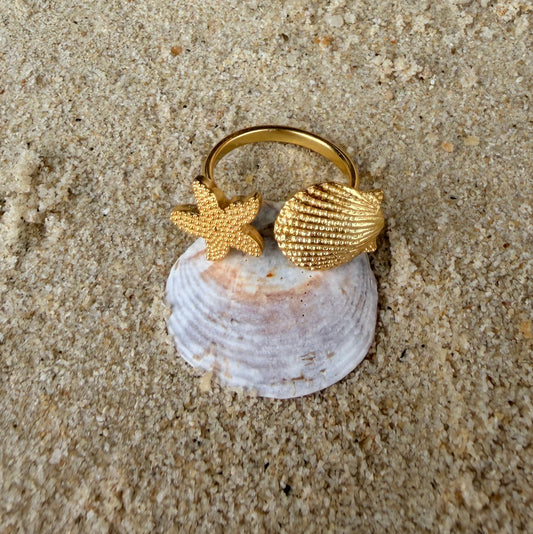 Gold ring with shell and starfish design on a sandy surface with pearls and shells.