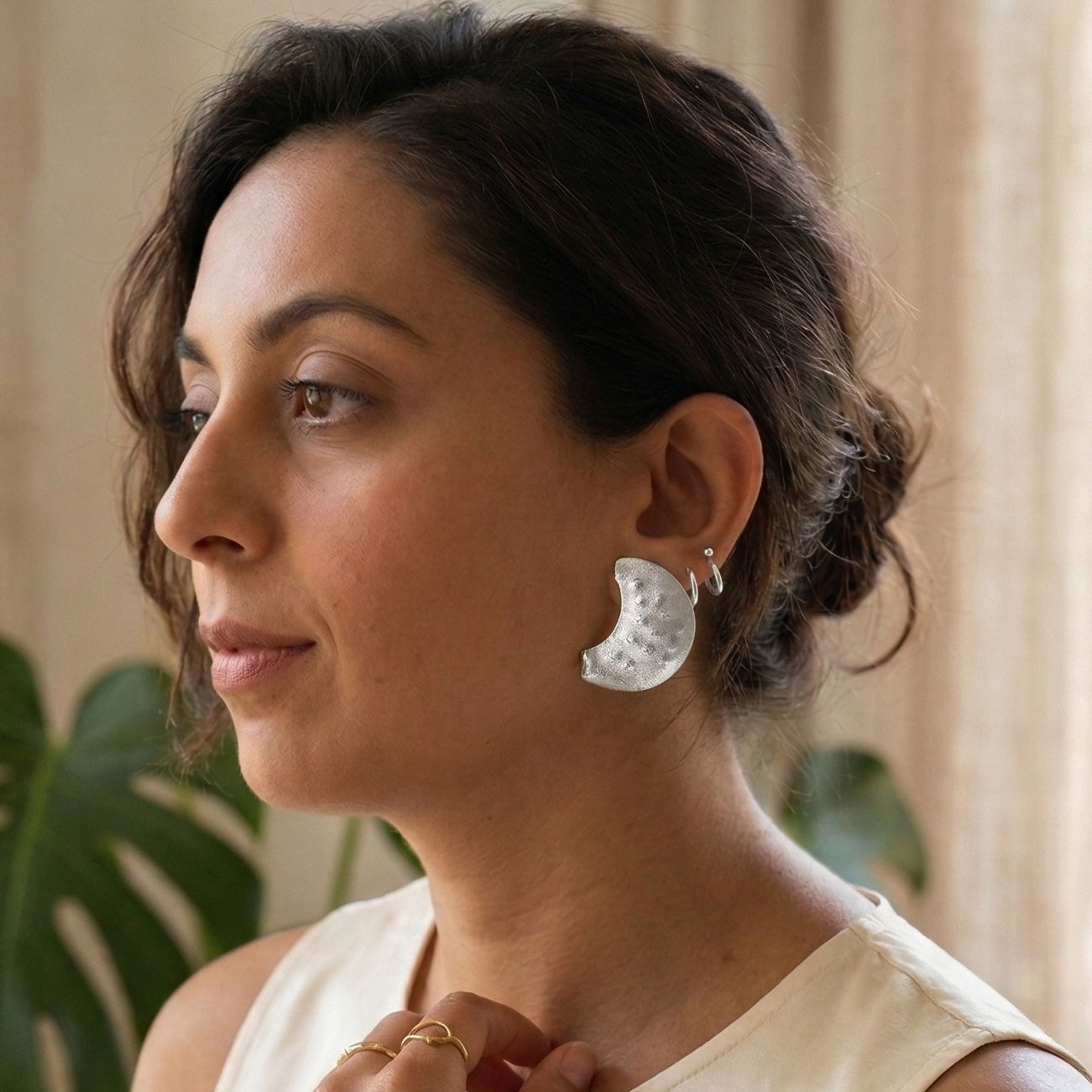 Woman wearing earrings and a necklace indoors with a plant and window in the background