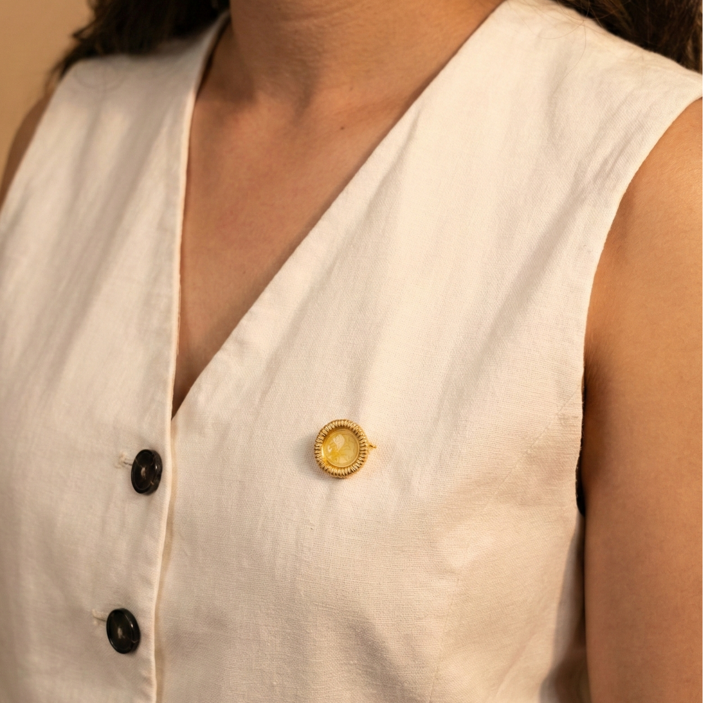 Close-up of a person wearing a white sleeveless top with a gold coin brooch on a beige background