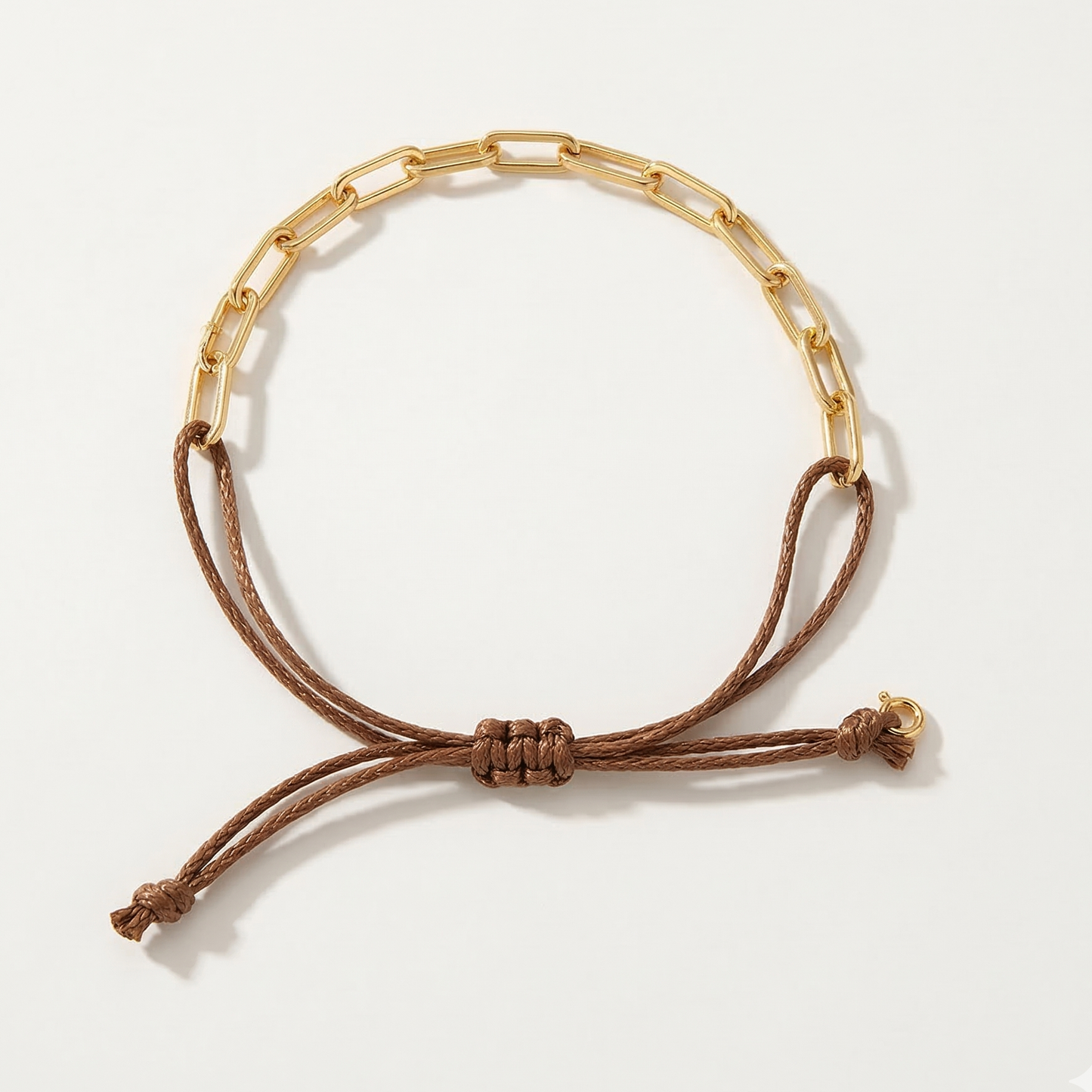 Gold paperclip chain bracelet with a brown cord on a white background