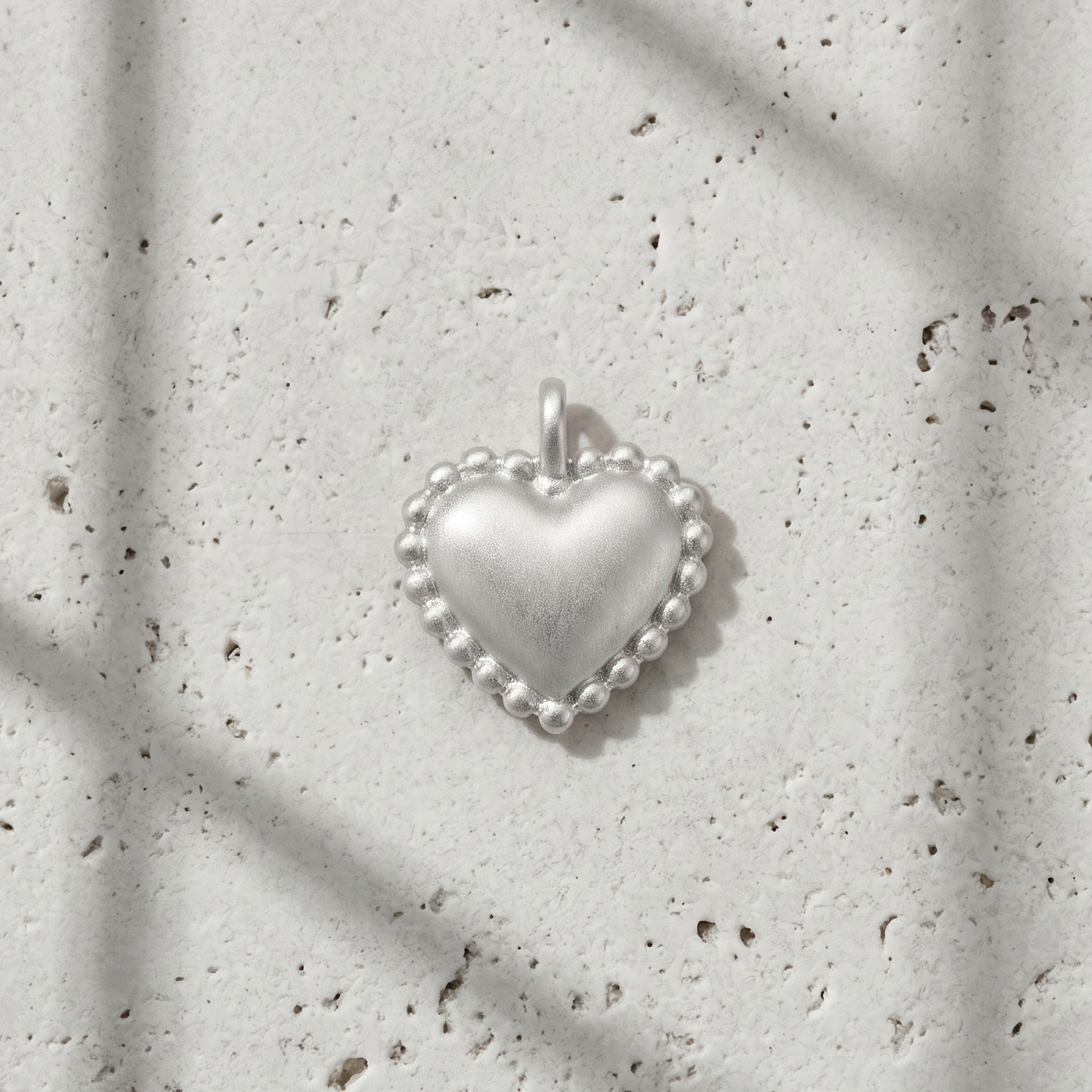 A small silver heart-shaped pendant with a beaded border and a top loop, photographed on a light textured surface with soft window-shadow lines.