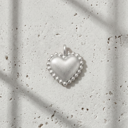 A small silver heart-shaped pendant with a beaded border and a top loop, photographed on a light textured surface with soft window-shadow lines.