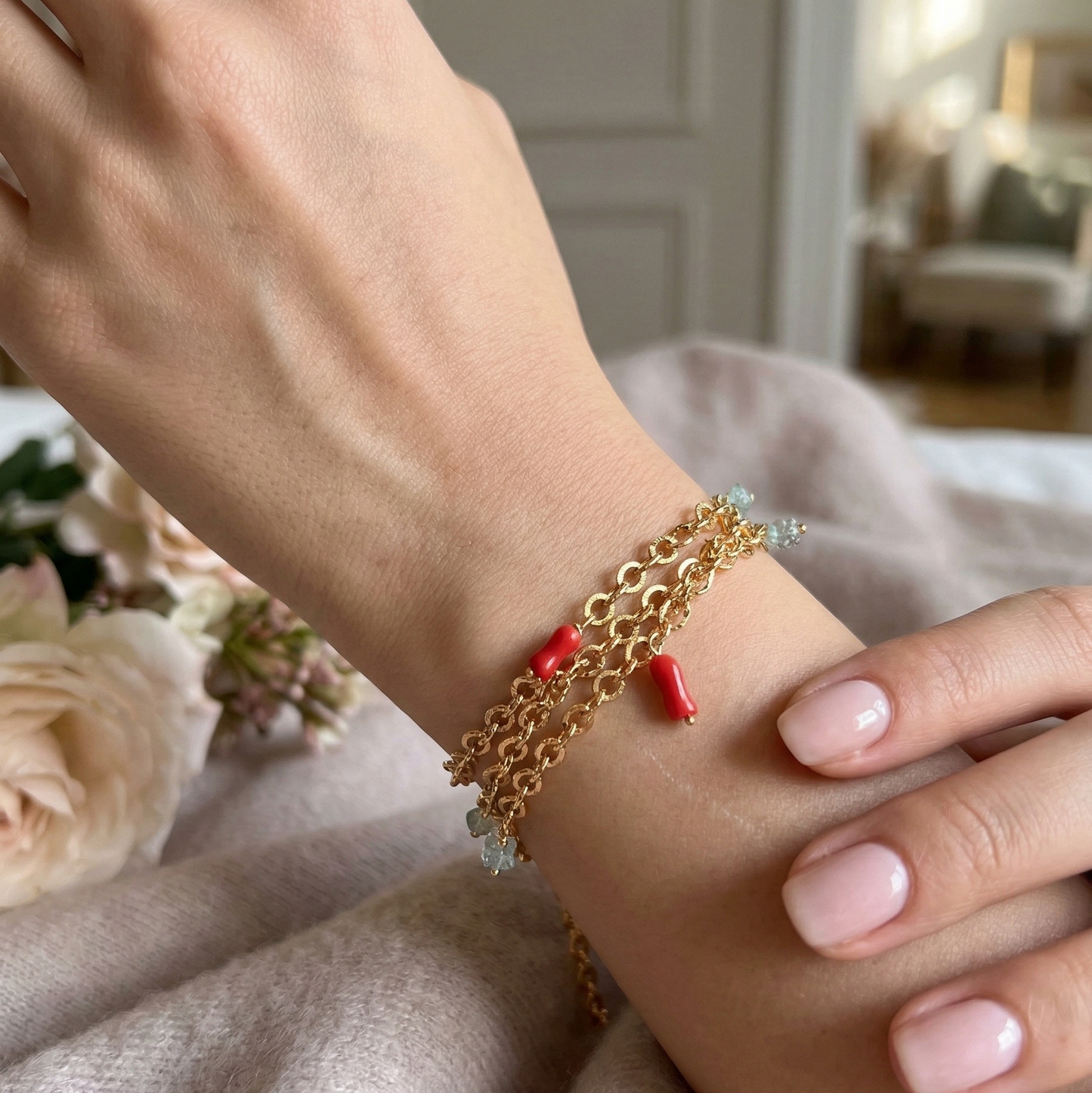Gold bracelet with red accents on a wrist, with flowers and a blurred indoor setting in the background.