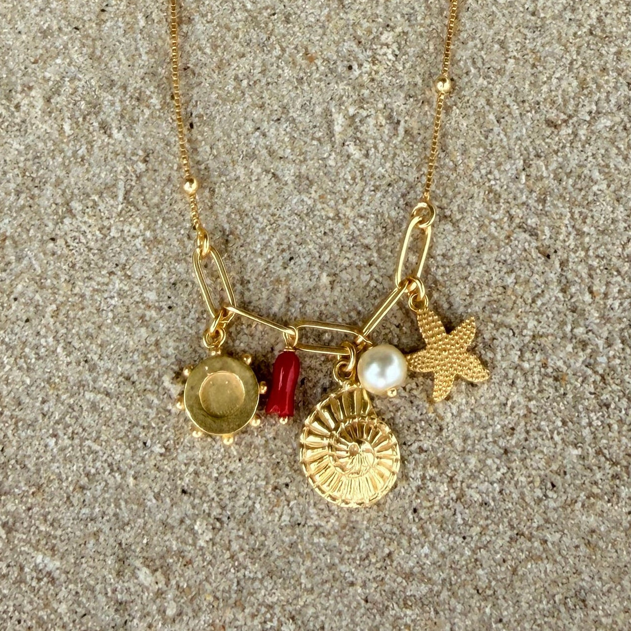 Gold necklace with charms pearl, coral, starfish, seashell on a sandy background