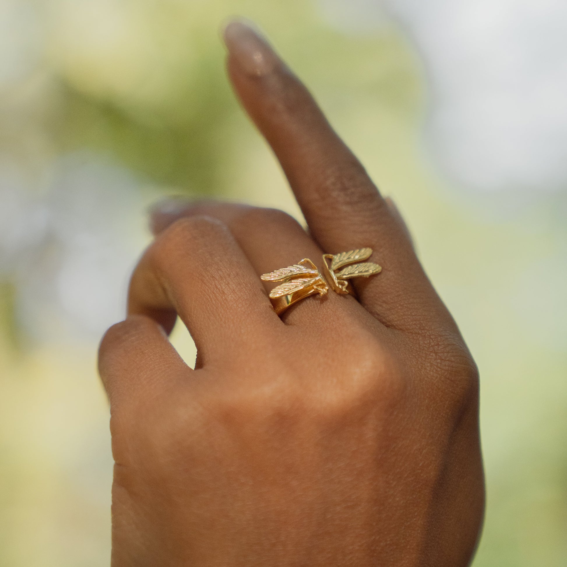 Hand wearing a gold dragonfly ring with a blurred natural background