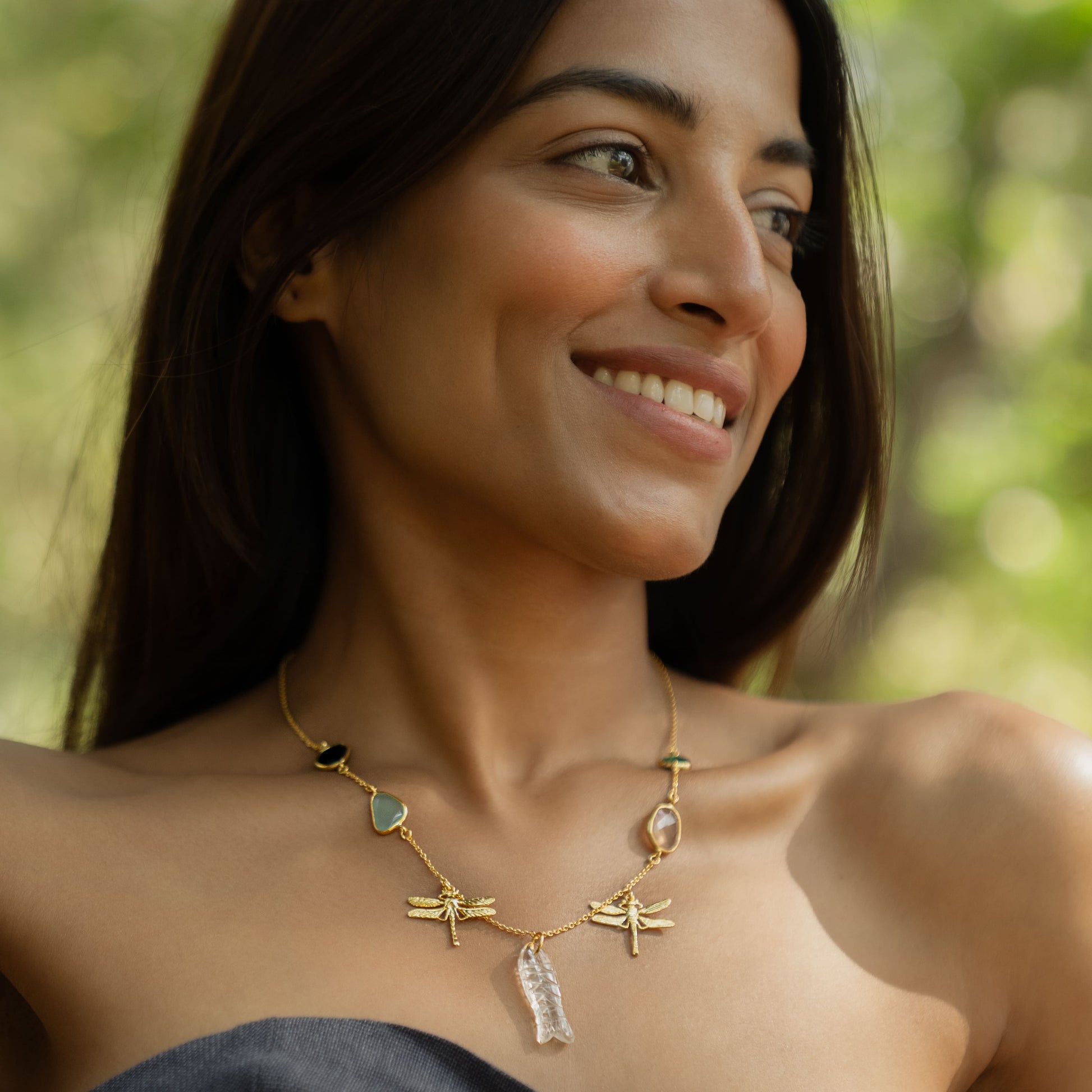 Woman wearing a gold necklace with green and blue stones, standing outdoors with greenery in the background.