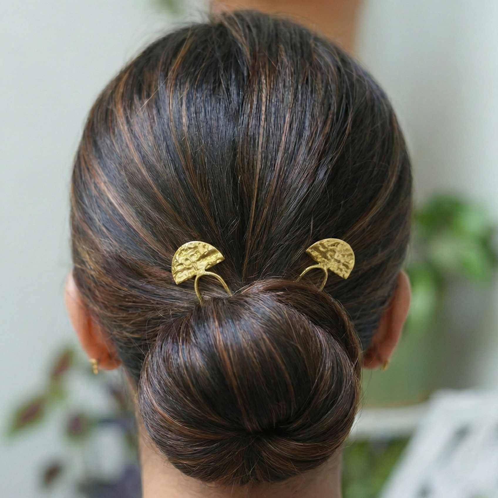 Person with a styled hair bun wearing gold hair accessories, with a blurred indoor background.
