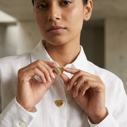 A woman wearing a white linen button-up shirt holds a small gold fan shaped brooch near her collarbone, with another matching fan shaped button cover.