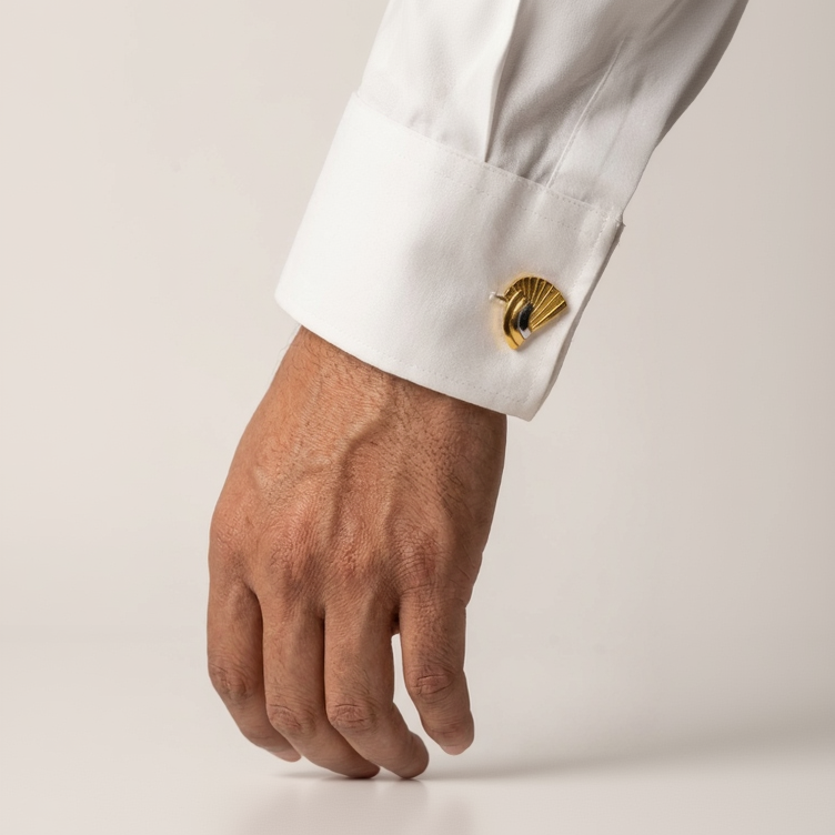 A close-up of a person's hand wearing a white dress shirt cuff with a gold-toned fan-shaped cufflink, set against a plain light background.