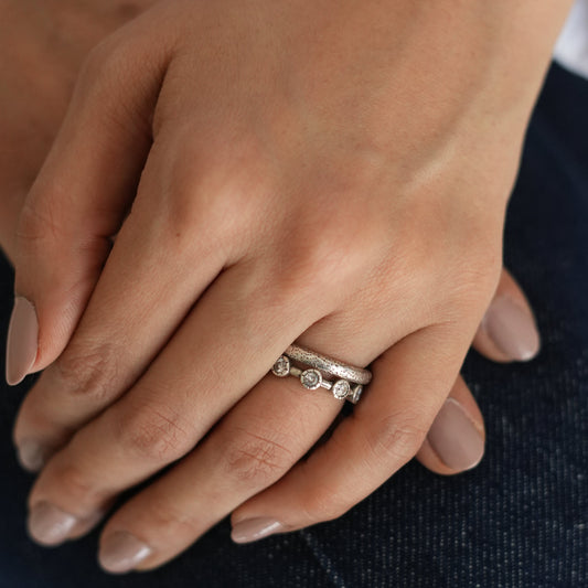 A close-up image of a hand wearing a silver stack ring with hammer texture and a small circular detail, possibly a gemstone or embellishment.