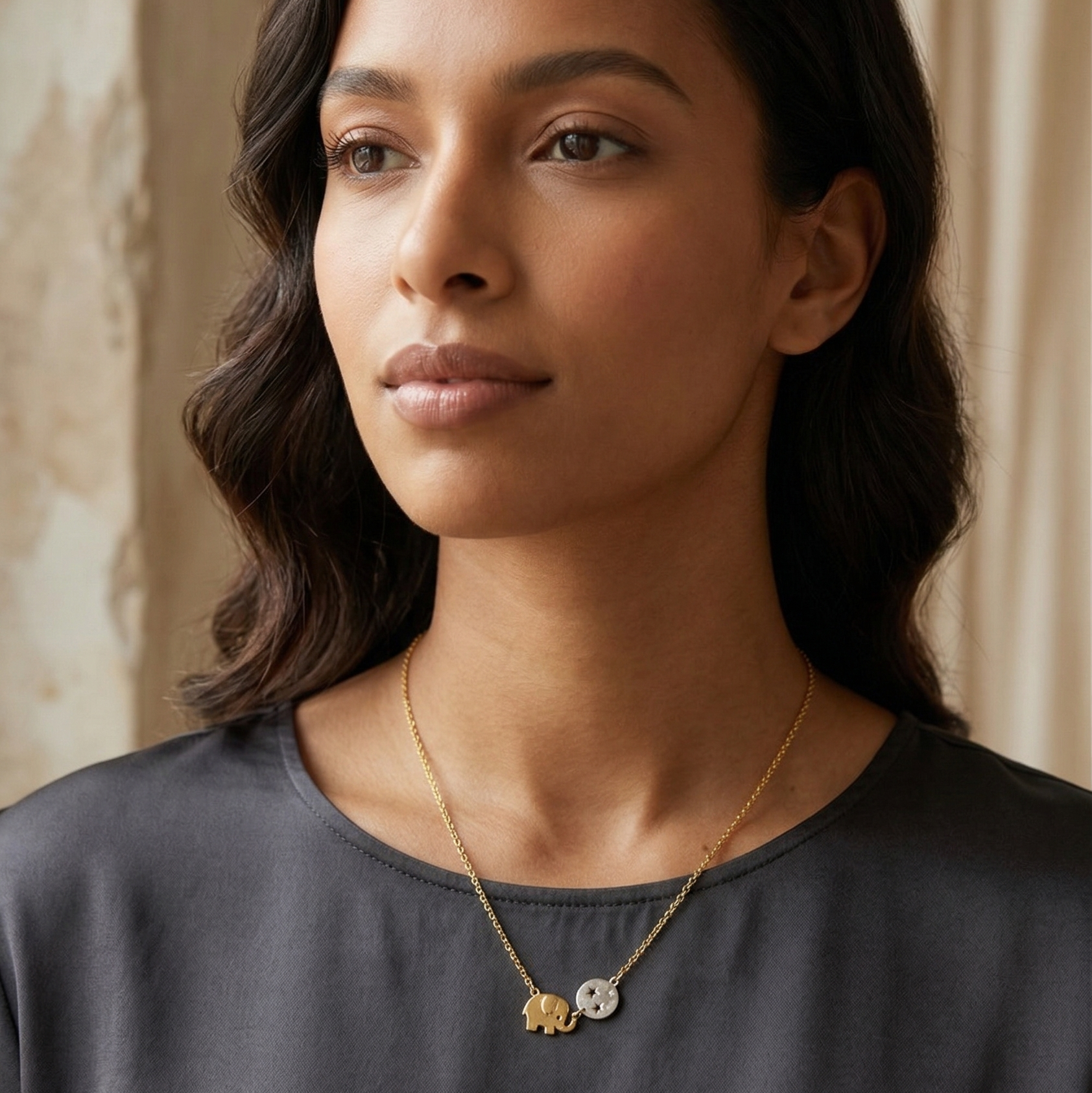 Woman wearing a gold necklace with small elephant and ball pendant against a neutral background