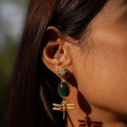Close-up of a person wearing gold earrings with green stones and dragonfly designs.