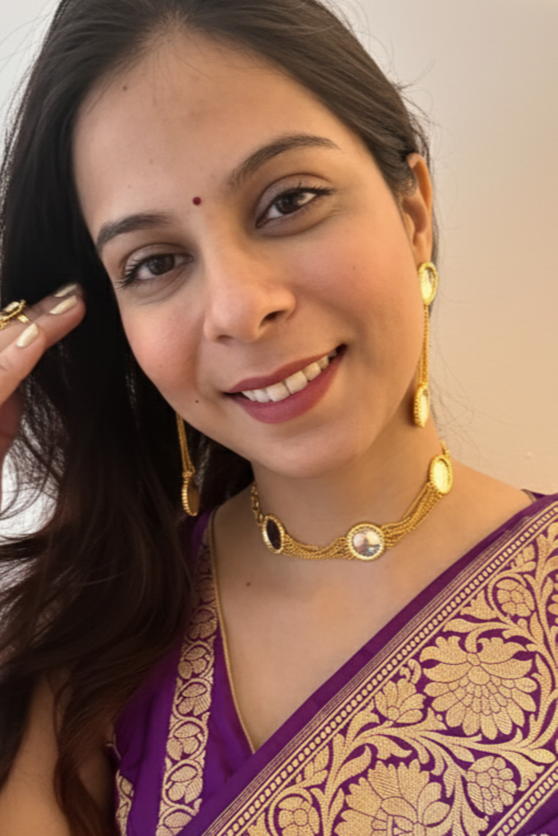 Woman wearing a purple saree with gold patterns, holding flowers, against a plain background