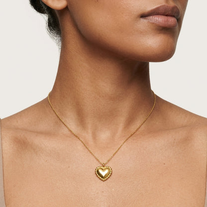A close-up of a model wearing a delicate gold chain necklace with a small heart-shaped pendant, shown against a neutral light beige background.