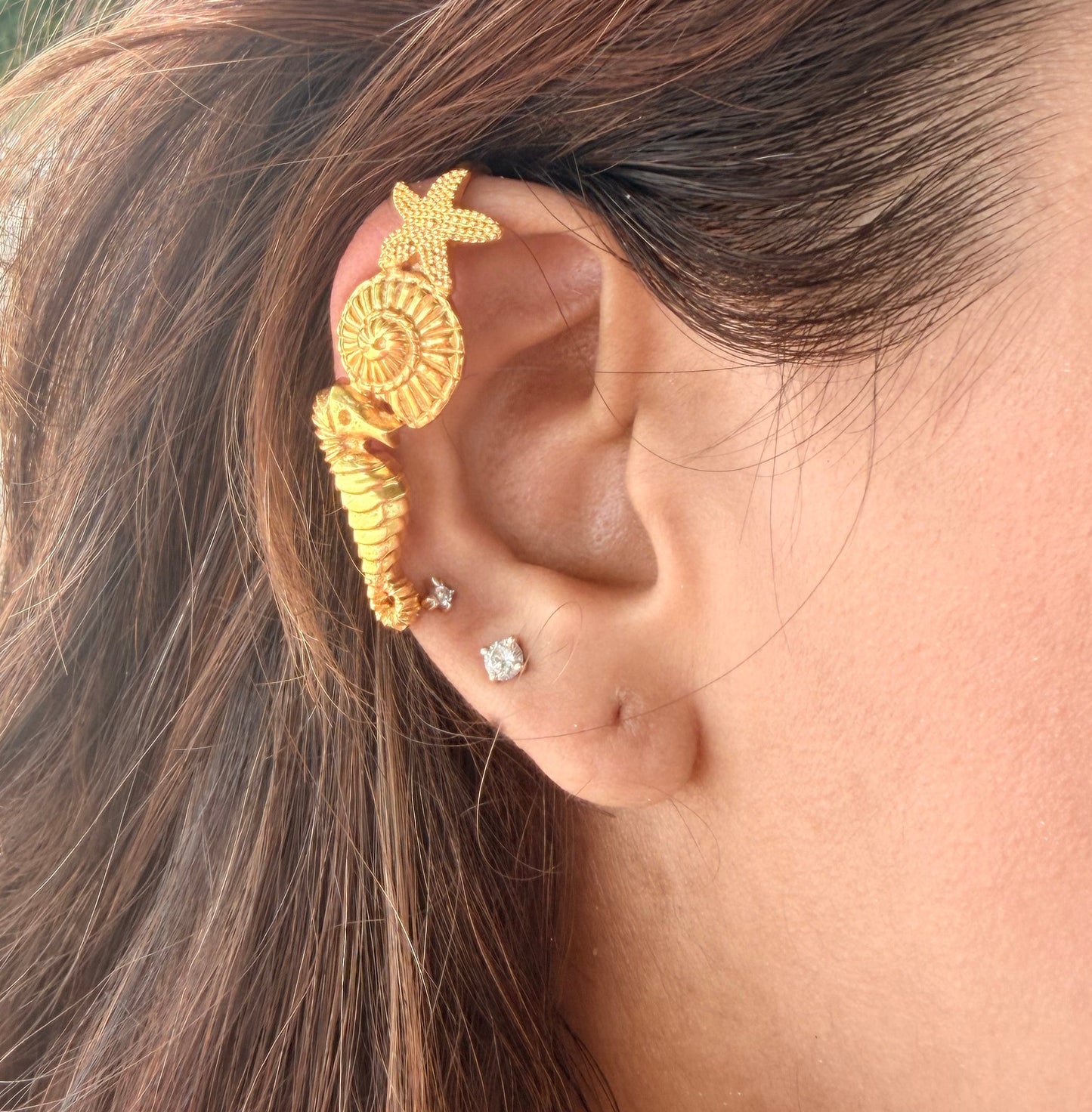 Gold seahorse and starfish-shaped earcuff on a sandy background with shells and pearls.