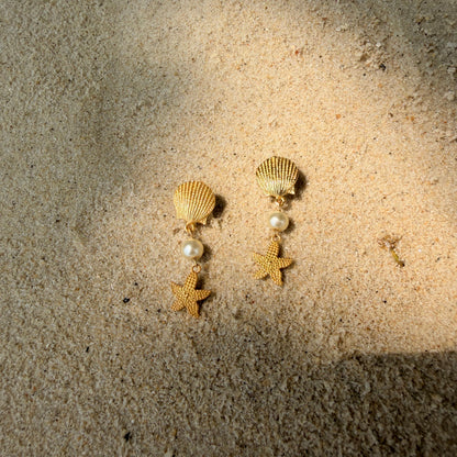 Gold starfish and pearl earrings on a sandy background with shells and pearls.