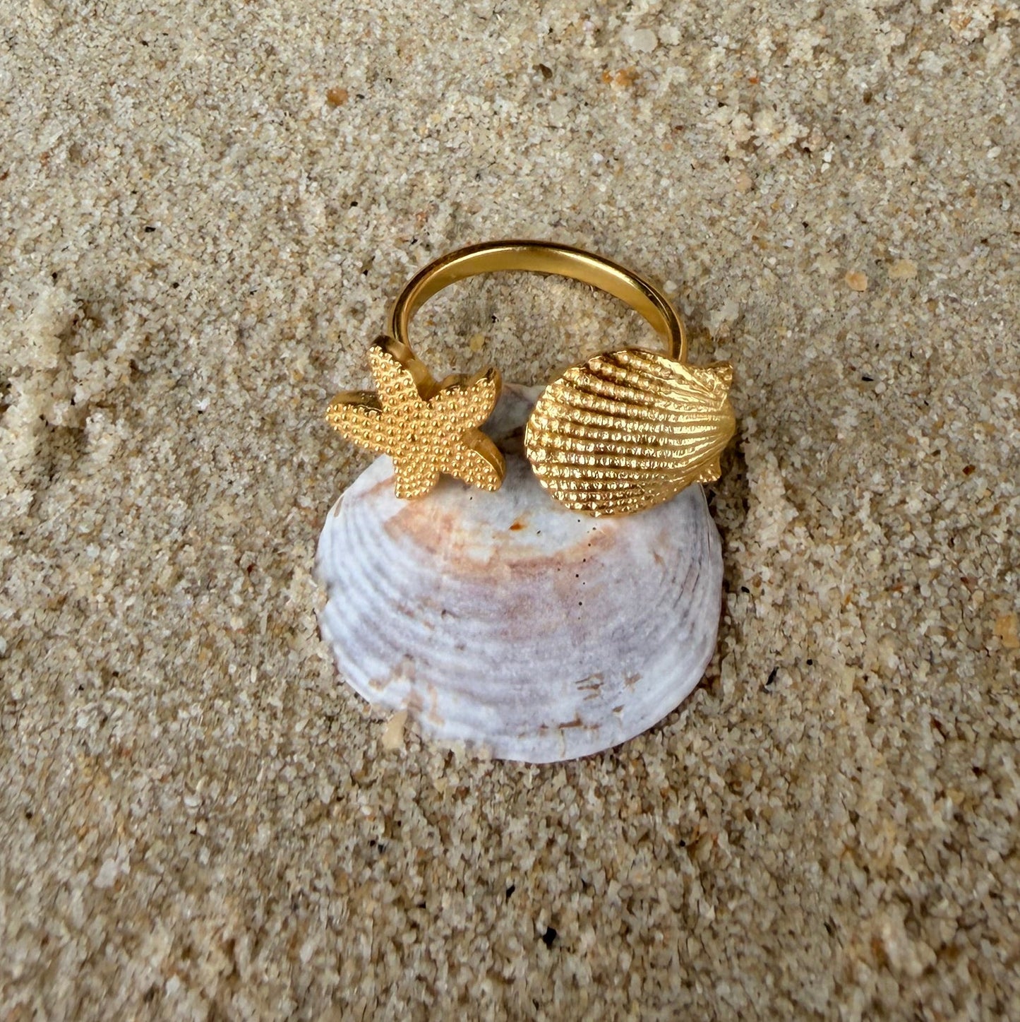 Gold ring with shell and starfish design on a sandy surface with pearls and shells.