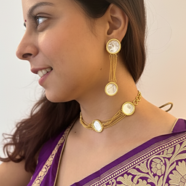 Woman wearing a purple saree with gold patterns, gold jewelry, and holding white flowers.