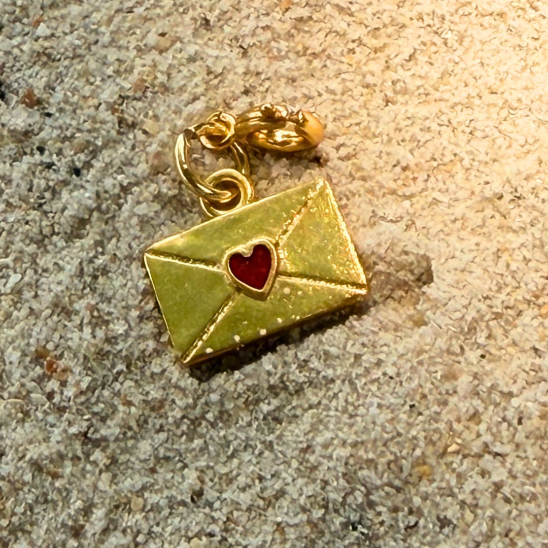 Gold charm shaped like an envelope with a red enamel heart on a textured surface