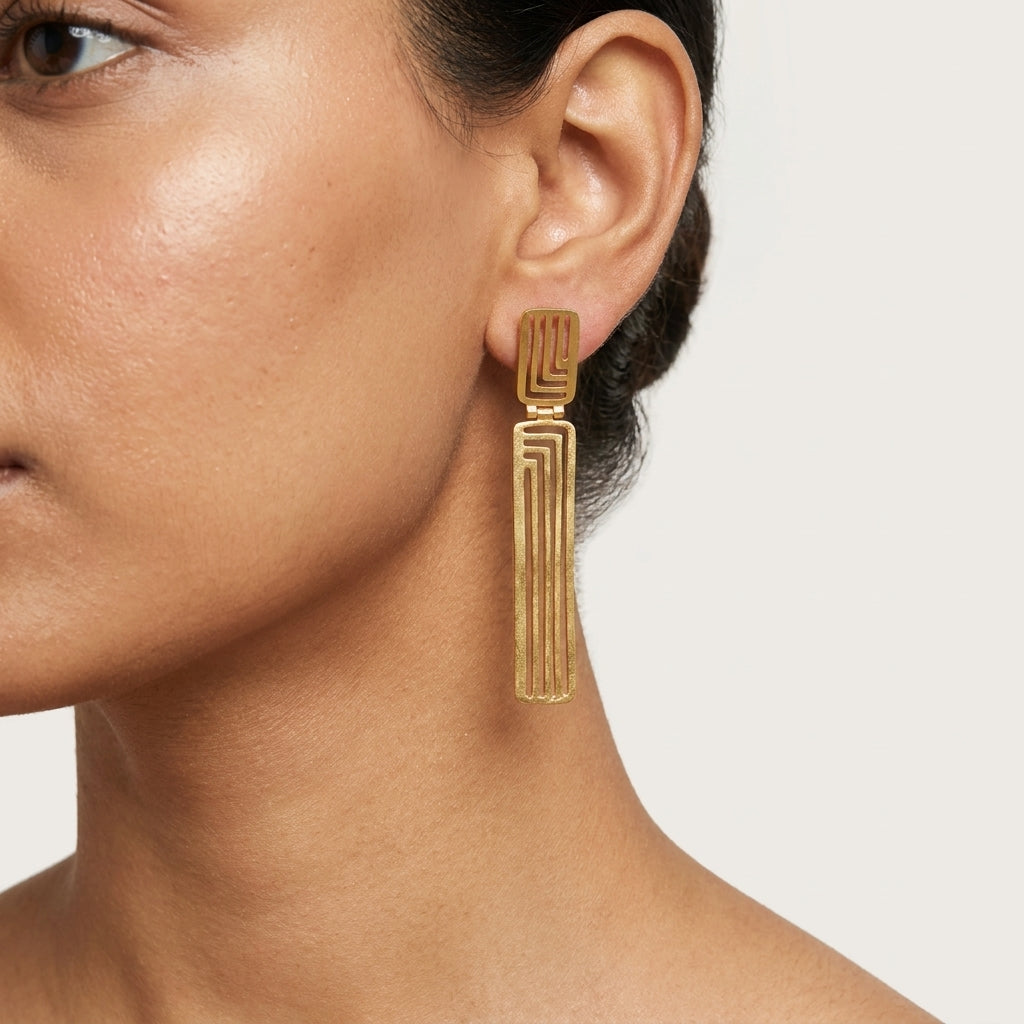 A close-up of a model's ear and neck wearing a single geometric gold dangle earring with a textured, ribbed vertical design.