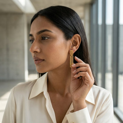A woman wearing a cream-colored collared blouse and a gold geometric drop earring, posing in a modern, softly lit office environment.