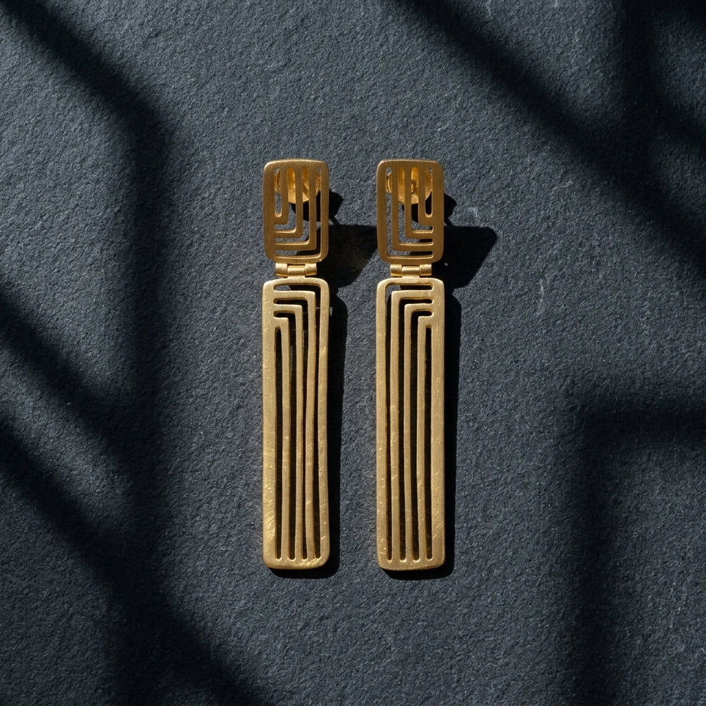 A pair of gold geometric drop earrings with a textured finish, displayed on a dark textured surface with soft shadow overlays.