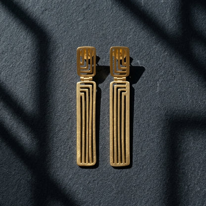 A pair of gold geometric drop earrings with a textured finish, displayed on a dark textured surface with soft shadow overlays.