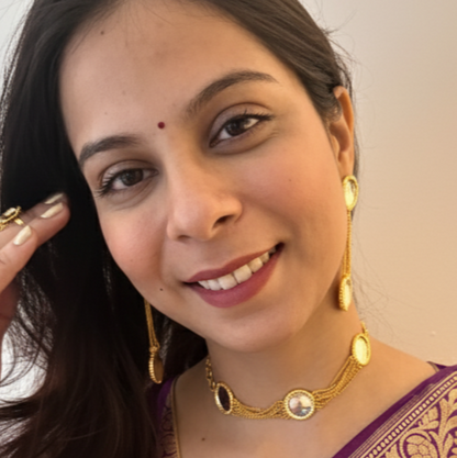 Woman wearing a purple saree with gold patterns, holding flowers, against a plain background