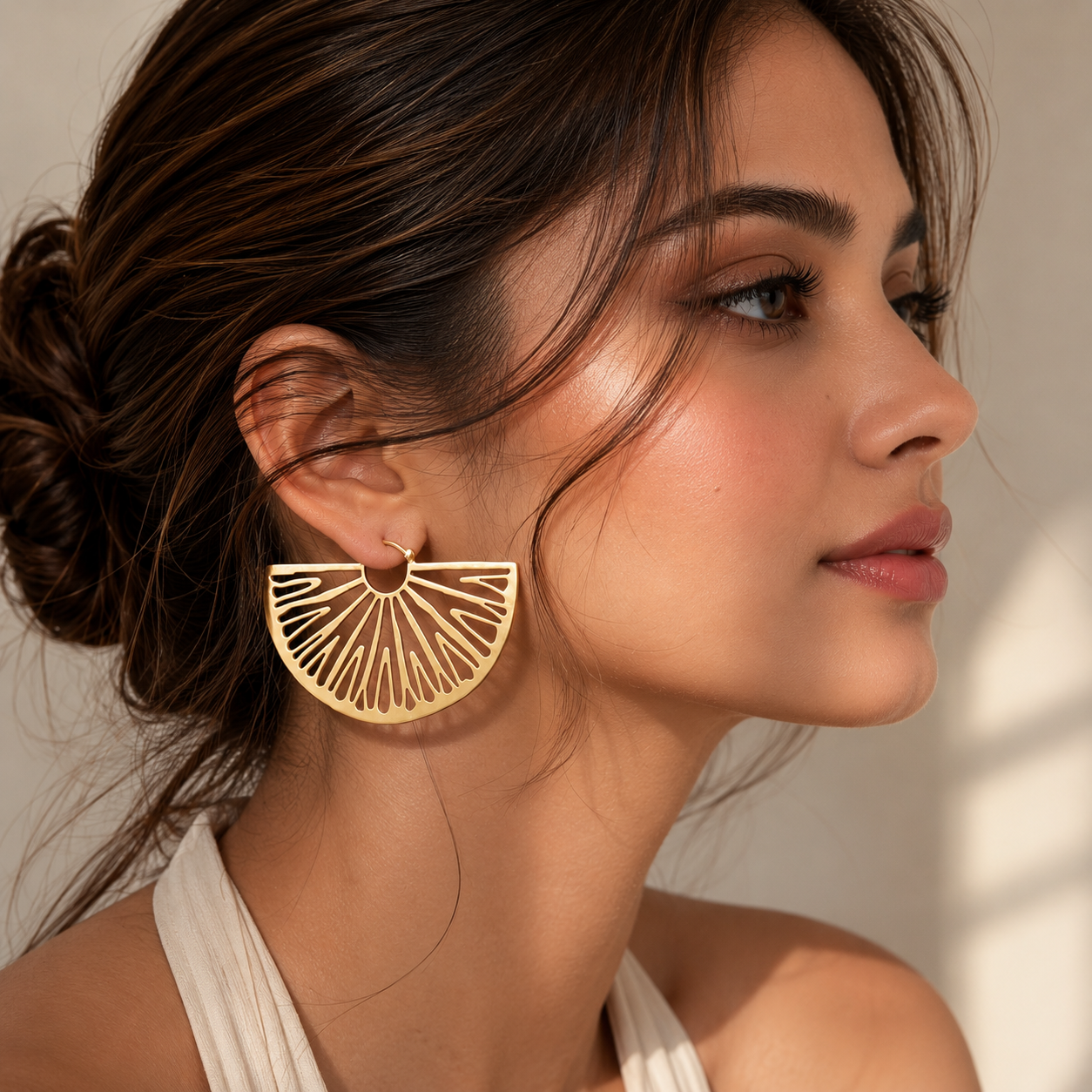 A woman in profile wearing a large gold geometric fan-shaped earring with a small gold hoop and drop stud, photographed against a softly lit neutral background.