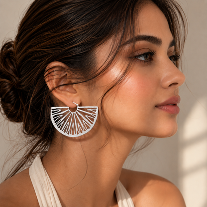 A woman in profile wearing a large silver fan-shaped dangle earring with a circular cutout design, set against a warm beige background with soft window shadows.