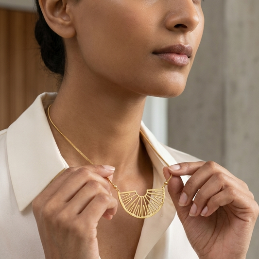 A woman wearing a gold geometric fan-shaped pendant necklace, holding the pendant with both hands against a soft-focus indoor background.