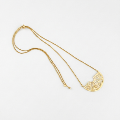 A gold-toned necklace featuring a delicate chain and a semi-circular fan-shaped pendant with radiating linear details, displayed on a clean white background.