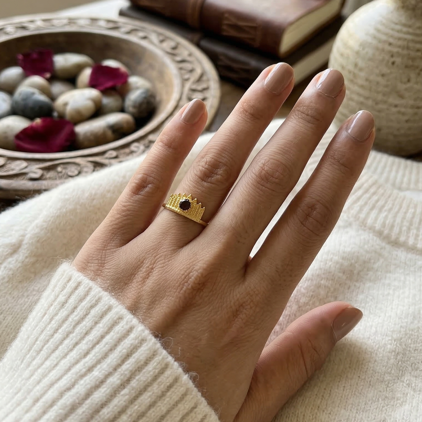 Hand wearing a gold ring with a garnet stone, set against a cozy indoor background.