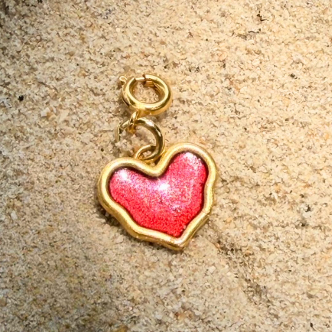 Heart-shaped charm with red enamel center and gold border on a beige surface