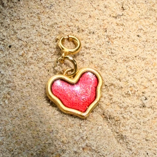 Heart-shaped charm with red enamel center and gold border on a beige surface