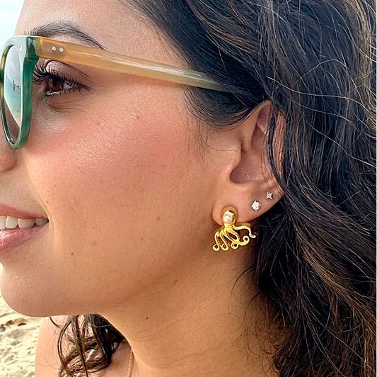 Gold octopus stud earrings with freshwater pearl accents on a textured peach surface.